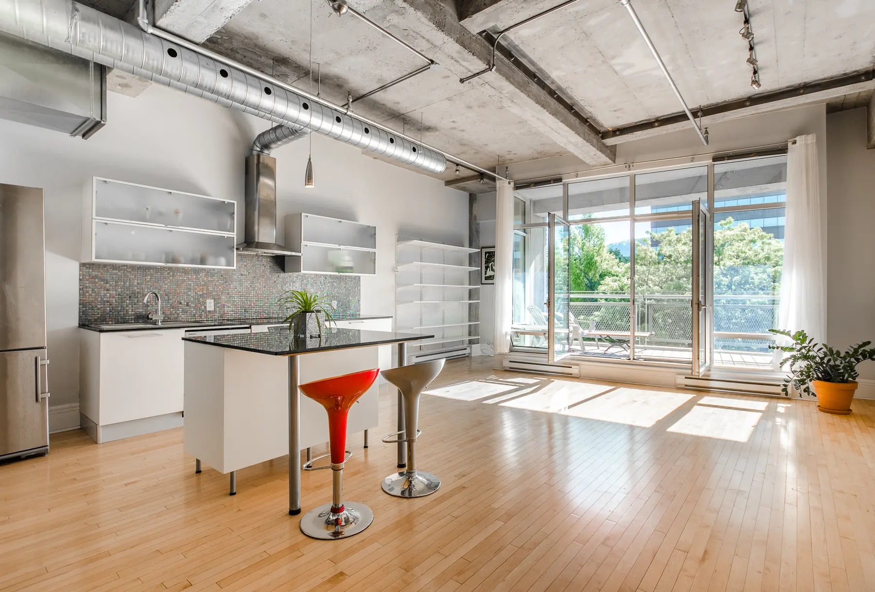 Salon ouvert d'un loft : plafond de béton, conduit d'aération apparent, mur de fenêtres donnant sur un balcon